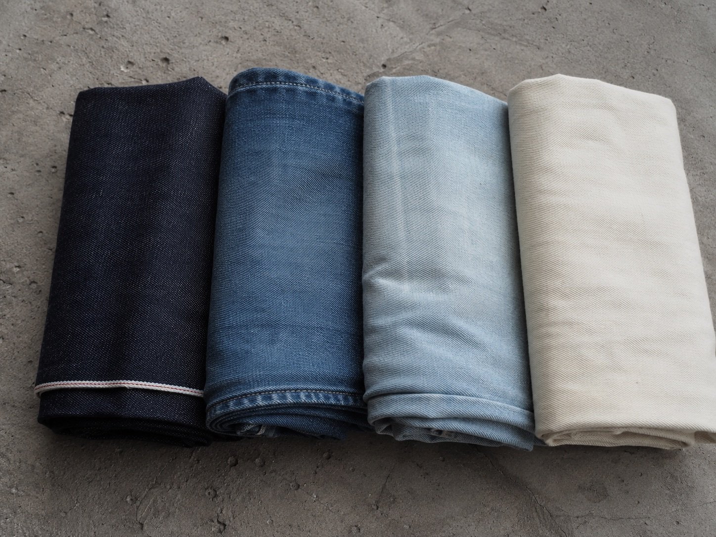 Four folded lengths of our denim, from raw selvedge indigo through mid and light washes to undyed